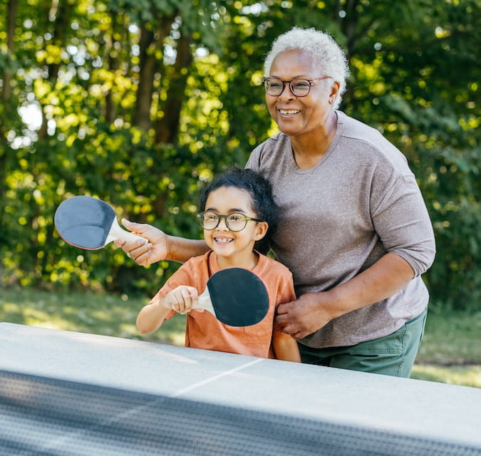 Photo of a grandmother and granddaughter playing ping pong outdoors.